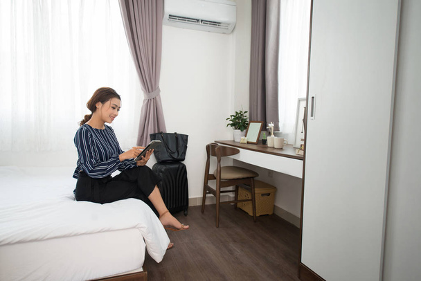 Χαρούμενη επιχείρηση κυρία σε μπλούζα κάθεται στο κρεβάτι και χρησιμοποιώντας το ψηφιακό δισκίο, ενώ προβολή online πληροφορίες στο δωμάτιο του ξενοδοχείου, βαλίτσα και τσάντα τοποθετημένο κοντά στον τοίχο - Φωτογραφία, εικόνα