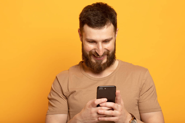 Close up shot portrait of young bearded man isolated on yellow background, looking agitated at display of her smartphone smiling and laughing happily, impressed by media content from web - Foto, Bild
