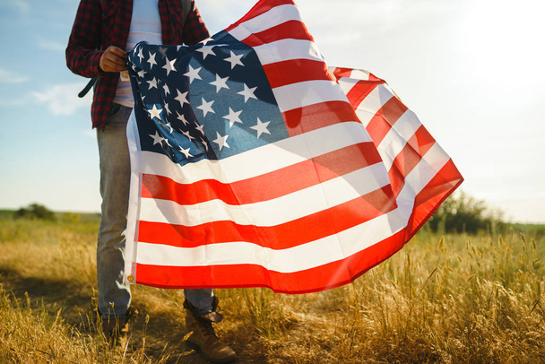 4 июля. Четвертое июля. Американец с национальным флагом. Американский флаг. День независимости. Патриотический праздник. На человеке шляпа, рюкзак, рубашка и джинсы. Закат света
.  - Фото, изображение