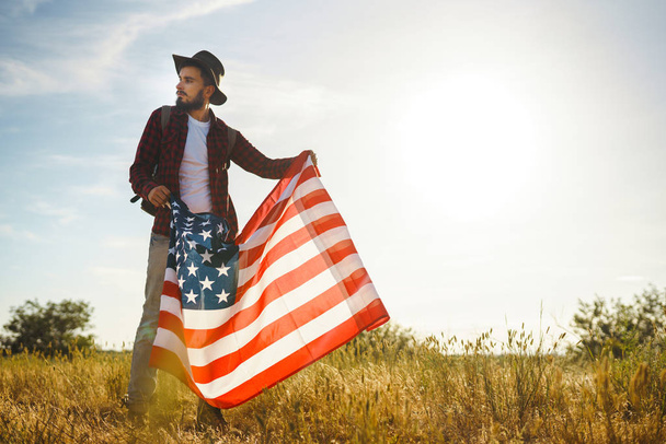 4. července. Čtvrtého července. Američan s národní vlajkou. Americký praporek. Den nezávislosti. Vlastenecká dovolená. Ten chlap nosí klobouk, batoh, košili a džíny. Nádherné světlo slunce.  - Fotografie, Obrázek