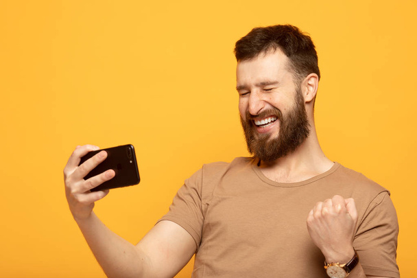 Portrait of an excited man standing over yellow background, holding mobile phone, celebrating - Foto, Imagen