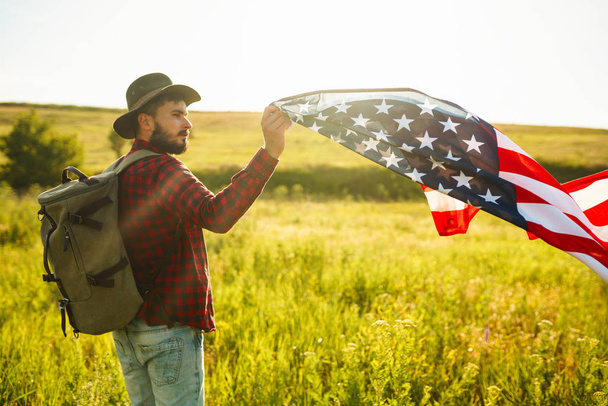 4th of July. Fourth of July. American with the national flag. American Flag. Independence Day. Patriotic holiday. The man is wearing a hat, a backpack, a shirt and jeans. Beautiful sunset light.  - Foto, afbeelding