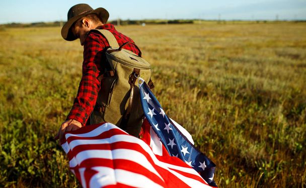 4th of July. Fourth of July. American with the national flag. American Flag. Independence Day. Patriotic holiday. The man is wearing a hat, a backpack, a shirt and jeans. Beautiful sunset light.  - Φωτογραφία, εικόνα