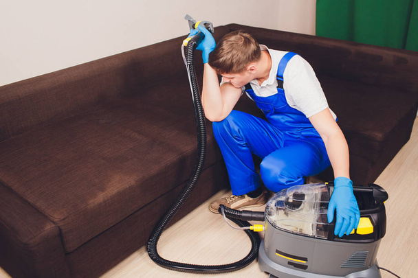 Tired young man sitting on sofa in living room. extractor, couch couch, chemistry, washing, cleaning. working blue uniform - Photo, Image