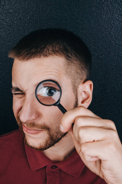 a young man with a beard looks through a magnifying glass. Portrait of a guy with a big eye on a black background. investigation, survey - Foto, afbeelding