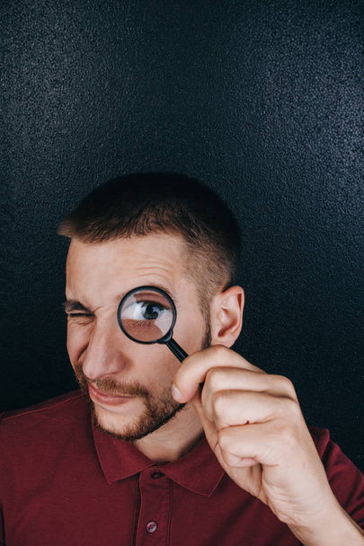 a young man with a beard looks through a magnifying glass. Portrait of a guy with a big eye on a black background. investigation, survey - Фото, изображение