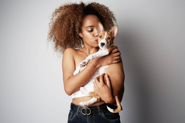 Handsome black african female with afro hair hugging and kissing her puppy basenji dog. Love between dog and owner. On white background - Foto, afbeelding