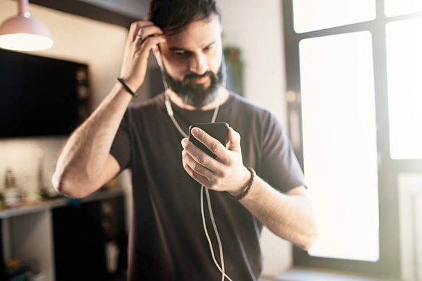 Handsome bearded guy wearing casual grey t-shirt listening music in earphones, checking social networks on smartphone having happy at home - Photo, Image