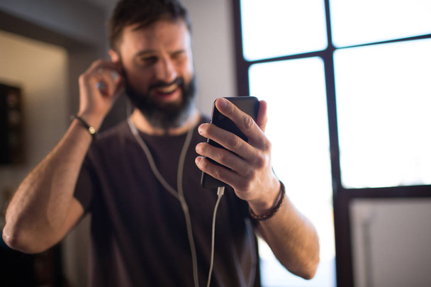 Young bearded guy wearing casual grey t-shirt listening music in earphones, checking social networks on smartphone having happy at home - Foto, Bild
