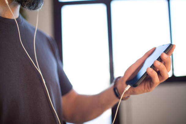 Tipo barbudo con camiseta gris casual escuchando música en los auriculares, comprobando las redes sociales en el teléfono móvil en casa
 - Foto, Imagen