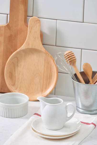 Simple rustic kitchenware against white wooden wall: rough ceramic pot with wooden cooking utensil set, stacks of ceramic bowls, jug and wooden trays. - Photo, Image