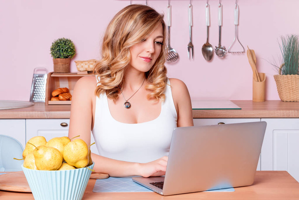 Χαμογελαστή νεαρή γυναίκα χρησιμοποιώντας φορητό υπολογιστή στην κουζίνα στο σπίτι. Ξανθιά γυναίκα εργάζεται στον υπολογιστή, ελεύθερος επαγγελματίας ή blogger που εργάζονται στο σπίτι - Φωτογραφία, εικόνα