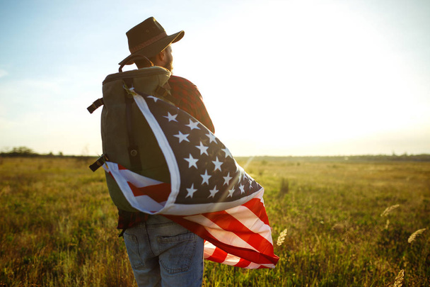 4th of July. Fourth of July. American with the national flag. American Flag. Independence Day. Patriotic holiday. The man is wearing a hat, a backpack, a shirt and jeans. Beautiful sunset light.  - Фото, изображение