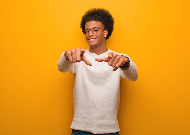 オレンジ色の壁の上に若いアフリカ系アメリカ人の男陽気で笑顔を前に指して - 写真・画像