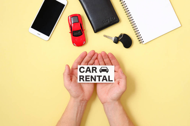 Car rental concept. Human hands holding text sign "CAR RENTAL", smartphone, driver license, toy car and automobile key on yellow background. Hire a car, renting service. Top view, flat lay composition - Photo, image