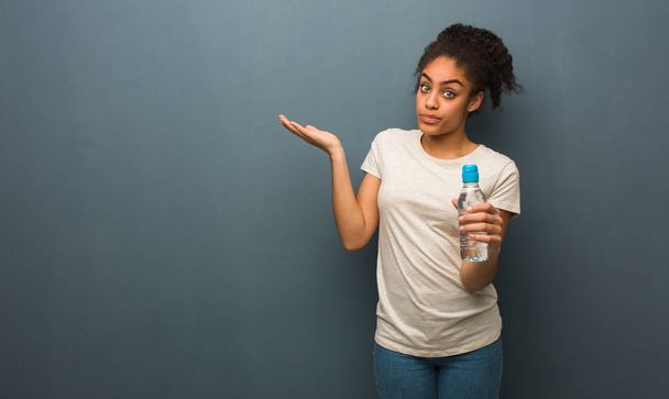 Νεαρή μαύρη γυναίκα μπερδεμένη και αμφίβολη. Κρατάει ένα μπουκάλι νερό.. - Φωτογραφία, εικόνα