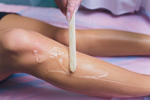The beautician is preparing for depilation and applying the cream with wax stick on the beautiful female legs. Beauty salon. - Фото, зображення