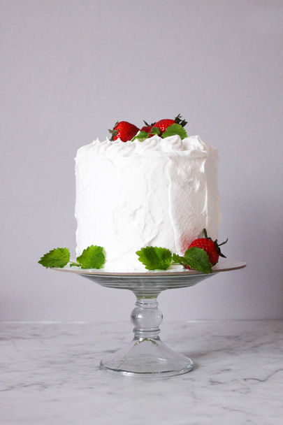 Large white delicious cake with cream and decorated with strawberries and mint on a transparent stand on a gray background   - Foto, Bild