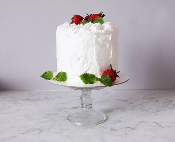 Large white delicious cake with cream and decorated with strawberries and mint on a transparent stand on a gray background   - Foto, afbeelding