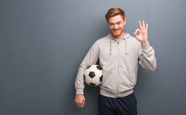 Молодой рыжеволосый фитнес-человек веселый и уверенный в себе делает хорошо жест. Он держит футбольный мяч.
. - Фото, изображение
