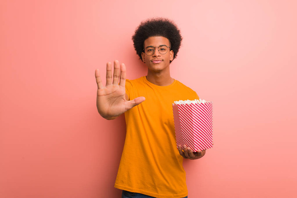 Young african american holding a popcorn bucket putting hand in front - Foto, imagen