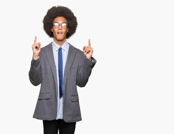 Jeune homme d'affaires afro-américain aux cheveux afro portant des lunettes étonné et surpris regardant vers le haut et pointant avec les doigts et les bras levés
. - Photo, image