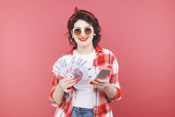 Портрет усміхненої дівчини, що тримає купу грошових купюр і дивиться на мобільний телефон ізольовано над рожевим
 - Фото, зображення