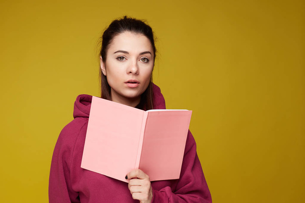 Teenage female in casual hoodie tries to learn important information in textbook, has concentrated serious face expression, isolated on yellow wall background. Education and exam preparation concept.  - Photo, Image