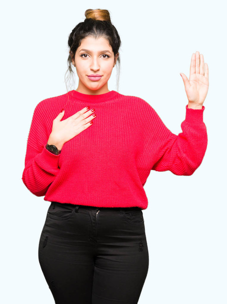 Giovane bella donna indossa maglione rosso e panino giurando con mano sul petto e palmo aperto, facendo un giuramento di fedeltà
 - Foto, immagini