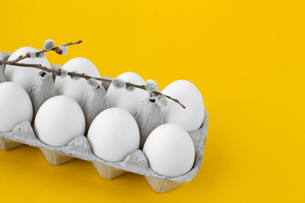 White chicken eggs in an open cardboard box on a yellow background. A willow twig lies on top. Healthy organic food and diet concept. Easter theme - Foto, Imagem