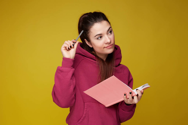 Η διέκφραστη νεαρή γυναίκα συγγραφέας κάνει παραστάσεις για νέες ιδέες, κρατά ροζ σημειωματάριο και μολύβι κοντά στο στόμα, έχει στοχαστική έκφραση, φοράει λιλά φούτερ, απομονωμένο σε κίτρινο φόντο με χώρο αντιγραφής στα δεξιά. - Φωτογραφία, εικόνα