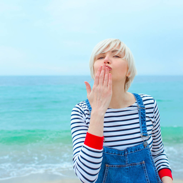 bella giovane donna caucasica bionda in vacanza in camicetta a righe, scarpe da ginnastica e denim nel complesso saltando sulla spiaggia dallo straordinario sfondo blu del mare
 - Foto, immagini
