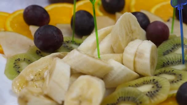 banai, naranjas, uvas, kiwi en rodajas, primer plano. Plato de fruta fresca en una mesa de comedor festiva. Brochetas de frutas rebanadas surtidas en un plato
. - Metraje, vídeo