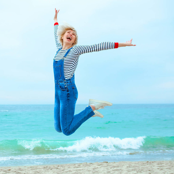mooie jonge blonde blanke vrouw op vakantie in gestreepte blouse, sneakers en denim overall op het strand door de blauwe zee achtergrond. Blij om aan zee te zijn - Foto, afbeelding