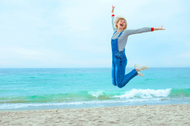 beautiful young blonde caucasian woman on vacation in striped blouse, sneakers and denim overall jumping on the beach by the amazing blue sea background - Photo, Image