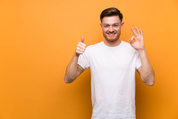 Redhead man over brown wall showing ok sign and thumb up gesture - Photo, Image