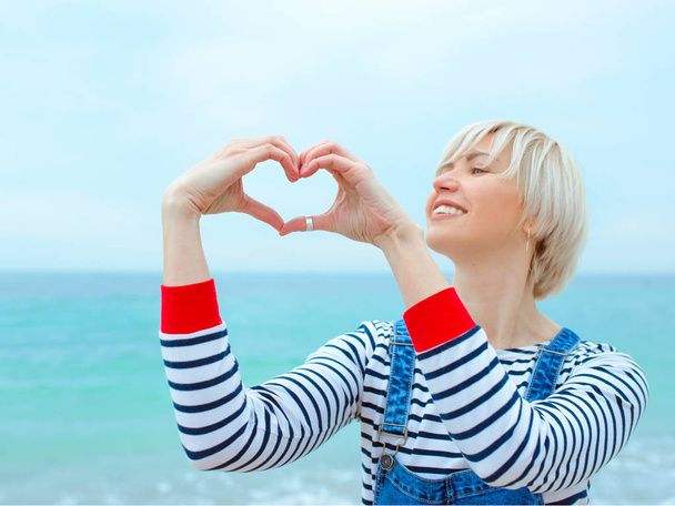 Beautiful young blonde caucasian woman with hands as love sign on vacation. Woman in striped blouse, sneakers and denim overall on the beach by the amazing blue sea background. Happy to be at the sea - Photo, Image
