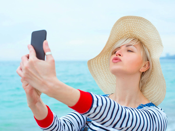 mooie jonge blonde Kaukasische vrouw op vakantie in strohoed, gestreepte blouse nad denim overalls maken selfie op smartphone door de verbazingwekkende blauwe zee achtergrond - Foto, afbeelding