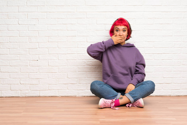 Молодая женщина с розовыми волосами сидит на полу покрывая рот руками
 - Фото, изображение