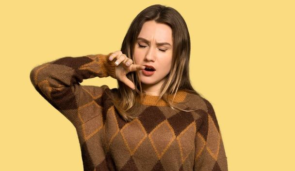 Teenager girl with brown sweater yawning and covering wide open mouth with hand over isolated yellow background - Photo, Image