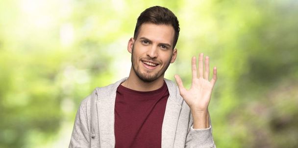 Ο άντρας με το πουλόβερ χαιρετά με το χέρι με καλή έκφραση σε ένα πάρκο - Φωτογραφία, εικόνα