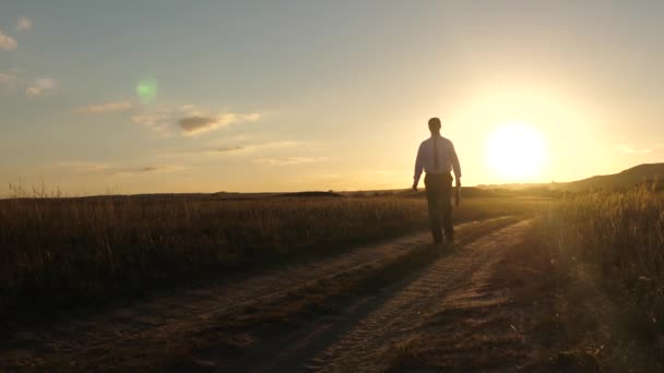 businessman in sunglasses goes down country road with a briefcase in his hand. agricultural business concept. The entrepreneur works in a rural area. farmer inspects the land at sunset. Slow motion. - Footage, Video