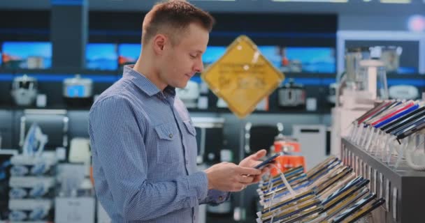 Giovane bell'uomo in camicia blu sceglie un nuovo telefono cellulare in un negozio di elettronica. Acquisto di tecnologia moderna
 - Filmati, video