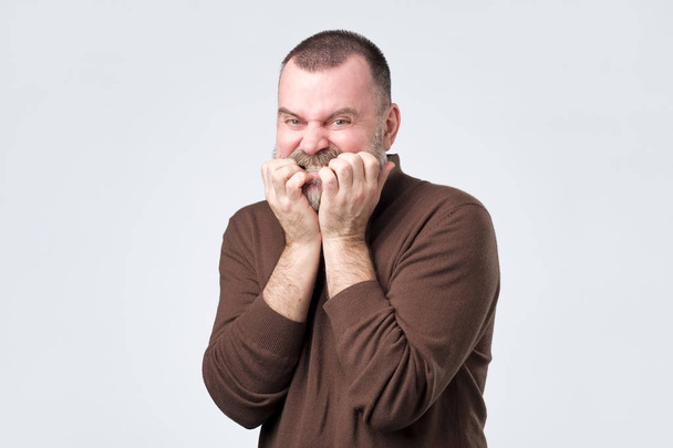 Человек с бородой в коричневой рубашке грызет гвозди в страхе
 - Фото, изображение