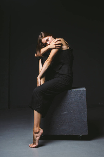 Социальная тема. молодая кавказская женщина в странной позе в одежде позирует на черном кубе, темном фоне, символизирует боль, страдания, обращение за помощью, защиту злого общества. Современное искусство, абстракция
 - Фото, изображение