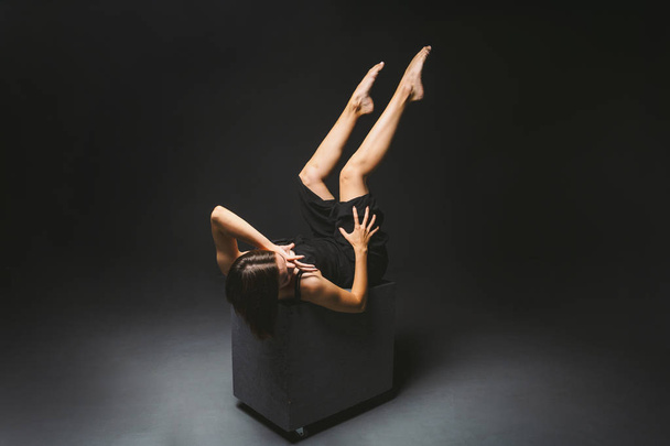 Social theme. young Caucasian woman in strange pose in dress posing on black cube, dark background, symbolizes pain, suffering, seeking help, protection of an evil society. Modern art, abstraction - Photo, Image