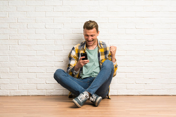Blond mężczyzna siedzi na podłodze z telefonem w pozycji zwycięstwa - Zdjęcie, obraz