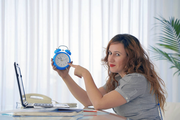 Жінка тримає годинник. Рудоволоса жінка тримає будильник у руці на робочому місці
. - Фото, зображення
