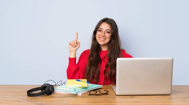 Adolescente estudiante chica estudiando en una mesa mostrando y levantando un dedo en señal de la mejor
 - Foto, Imagen