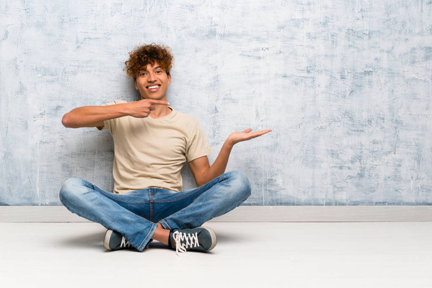 Fiatal afro-amerikai férfi ül a padlón, a tenyerén képzeletbeli másolópapírt tartva, hogy reklámot tegyen fel. - Fotó, kép